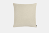 A square, light beige pillow with a soft texture is shown against a plain white background. A small black fabric tag is attached to one corner of the pillow.