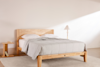 A minimalist bedroom with a light wood bed frame, a gray quilt, white pillows, a small wooden side table with books, and a floor lamp with a white shade against a plain off-white wall.