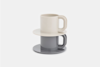 Two modern, stackable cups with matching saucers, one in white and one in gray, arranged in a minimalist style against a plain white background.