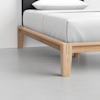 A close-up of a modern wooden bed frame with clean lines, light wood finish, and white bedding on a smooth, light gray floor. The image focuses on one corner of the bed.
