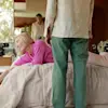 A woman in a pink sweater leans and smiles on a bed, reaching out. A man stands at the foot of the bed, seen from behind, and another man is visible in the background, looking toward them.