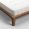 Close-up of a mattress with a white quilted cover on a wooden bed frame with clean lines and sturdy legs, set on a light gray floor.
