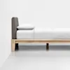 A minimalist bed with a wooden frame, gray upholstered headboard, and white bedding is positioned against a plain white wall in a bright, uncluttered room.