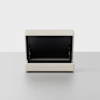 A beige, rectangular upholstered storage ottoman with its top lid partially open, revealing a black interior compartment. The ottoman sits on a smooth, light-colored surface against a plain white background.