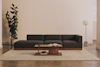 A minimalist living room with a dark gray sectional sofa, a low wooden coffee table holding an open book and cup, a beige rug, a tall plant, a floor lamp, and neutral-toned walls and flooring.