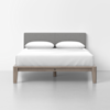 A modern bed with a light wood frame, white bedding, two white pillows, and a gray upholstered headboard, set against a plain white wall and light-colored floor.