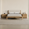 A minimalist bedroom features a wooden bed with a light upholstered headboard, white bedding, two matching wooden nightstands, a rug, a lamp, books, and a small vase with branches. The walls are light beige.