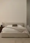 A neatly made bed with a beige upholstered frame, white bedding, a brown accent pillow, and a light beige throw blanket. A dark wooden nightstand with a white lamp and a small black object sits to the left.