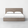 A wooden bed frame with a high headboard, dressed in white bedding and two white pillows, stands against a plain white wall on a light gray floor.