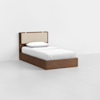 A single bed with a wooden frame, beige cushioned headboard, white sheets, and a white pillow, placed in a minimalist room with white walls and a light-colored floor.