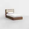 A single bed with a wooden frame, beige cushioned headboard, white sheets, and a white pillow, placed in a minimalist room with white walls and a light-colored floor.