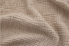 Close-up of beige, textured fabric with a crinkled, grid-like pattern. The material has soft folds and shadows, highlighting its three-dimensional surface and cozy appearance.