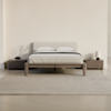 A minimalist bedroom with a bed featuring a light-colored upholstered headboard, two white pillows, gray bedding, wooden frame, two wooden nightstands, a lamp, books, and a decorative branch, all on a beige rug.
