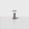 A small, modern wooden side table with a square top and a solid central base, set against a plain white background.