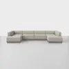A large, modern sectional sofa in a U-shape with light beige upholstery and low wooden legs, set against a plain white background and gray floor.