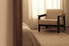 A beige armchair with wooden arms sits on a light carpet beside sheer curtains in a softly lit, minimalist room. Part of a bed covered with a textured blanket is visible in the foreground.