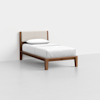 A single bed with a wooden frame and light upholstered headboard, dressed in white bedding with one pillow, placed in a minimalist room with light gray flooring and white walls.