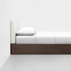 A minimalist bed with a padded beige headboard, white bedding, and a dark wooden frame, positioned against a plain white wall on a light gray floor.