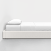 A minimalist bedroom with a low, modern white bed frame, neatly made with white sheets and a single white pillow, set against a plain white wall and light gray floor.