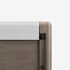 Close-up of the corner of a wooden crib or bed frame with a light gray fabric rail cover and rounded edges, against a plain white background.