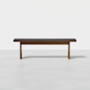 A minimalist wooden bench with a dark cushioned seat, shown against a plain white background. The bench has clean lines and rectangular legs.