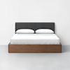 A modern bed with a dark gray upholstered headboard, white bedding, two pillows, and a wooden frame, set against a plain white wall and light gray floor.