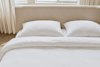 A neatly made bed with a beige upholstered headboard, two white pillows, and a white textured comforter, positioned in front of large windows with light-colored curtains.