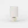 A modern table lamp with a cylindrical white fabric shade and a round, matte cream base featuring a small circular knob on the front, set against a plain white background.