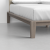 Close-up of a modern wooden bed frame with light brown legs and a neatly made bed with white sheets and pillows, placed on a light gray floor against a white wall.
