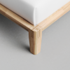 Close-up of a wooden bed frame corner with clean, modern lines and visible wood grain, showcasing precise joinery. A white mattress sits on top. The floor is light gray and smooth.