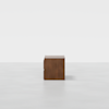 A small, square, wooden cabinet with two drawers sits on a plain, light gray floor against a white background.
