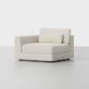 A modern, light beige upholstered armchair with a wide seat, low backrest, and one armrest on the left side, featuring a rectangular back cushion and a smaller lumbar cushion, set against a plain white background.