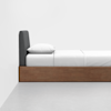 A minimalist bed with a wooden frame, a gray upholstered headboard, and white bedding with a single pillow, set against a plain white wall and light-colored floor.