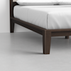 Close-up of the corner of a modern wooden bed frame with clean lines and dark finish, topped with a neatly made white mattress and bedding, set against a light grey floor and white wall.