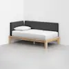 A minimalist daybed with a light wood frame, white mattress, single white pillow, and dark gray upholstered back and side cushions, placed in the corner of a plain, light-colored room.