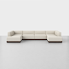 A large, modern, U-shaped sectional sofa with light beige upholstery and a low, dark base, positioned on a smooth, light gray floor against a plain white background.