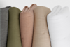 Five rolled-up textured fabrics are standing vertically side by side. The fabrics are colored dark gray, olive green, dusty pink, beige, and white, each with a crinkled, soft appearance.