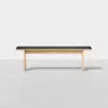 A minimalist wooden bench with light-colored legs and frame, topped with a flat black cushion, positioned against a plain white background.