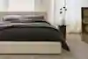 A modern bedroom with a low beige bed frame, dark bedding, two pillows, a gray rug, and a round wooden nightstand holding a vase with dried flowers, all set against large windows with sheer curtains.
