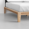 Close-up of a bed corner showing a minimalist light wood frame and legs, with a neatly made white mattress and bedding on top, set on a smooth, light gray floor.
