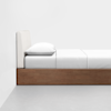 Side view of a minimalist bed with a wooden frame, white bedding, and a beige upholstered headboard, positioned against a plain white wall on a gray floor.