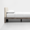 A minimalist bed with a light beige upholstered headboard, white bedding, and a simple wooden frame set against a plain white wall and floor.