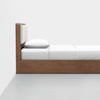 A side view of a wooden bed with a white mattress, white pillow, and white bedding, positioned against a plain white wall and on a light gray floor.