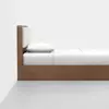 A side view of a wooden bed with a white mattress, white pillow, and white bedding, positioned against a plain white wall and on a light gray floor.