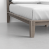 A close-up view of a wooden bed frame with light gray finish, white bedding, and a smooth gray floor in a minimalistic room.