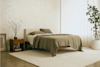 Minimalist bedroom with a wooden bed, beige bedding, and neutral decor. A small nightstand with a vase and framed art sits beside the bed. Large potted plant and sheer curtains complete the calm, airy space.
