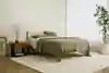 Minimalist bedroom with a wooden bed, beige bedding, and neutral decor. A small nightstand with a vase and framed art sits beside the bed. Large potted plant and sheer curtains complete the calm, airy space.