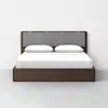 A modern bed with a dark wooden frame, a gray upholstered headboard, white bedding, and two white pillows, set against a plain white wall and light gray floor.