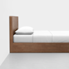 A minimalist wooden bed with a high headboard, made up with white sheets and a white pillow, positioned against a white wall on a light gray floor.
