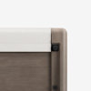 Close-up of the corner of a light wood bed frame with a white fabric-covered safety rail attached to the top edge, set against a plain white background.